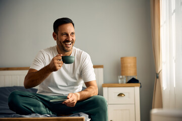 Happy man drinking coffee while relaxing on bed in morning.