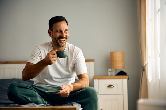 Happy man drinking coffee while relaxing on bed in morning.