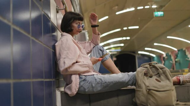 Young woman waiting for train at station and using her phone. watch schedule. Traveling by public transport.  Young girl in stylish clothes with backpack in subway. 