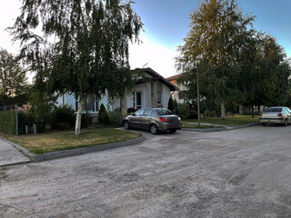 A quaint countryside house is set against a serene backdrop, featuring a parked car in the driveway. The evening light casts a warm glow on the well-maintained yard