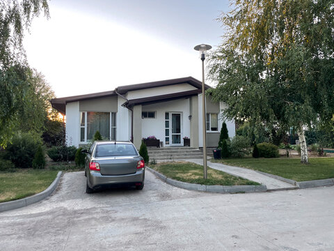A quaint countryside house is set against a serene backdrop, featuring a parked car in the driveway. The evening light casts a warm glow on the well-maintained yard