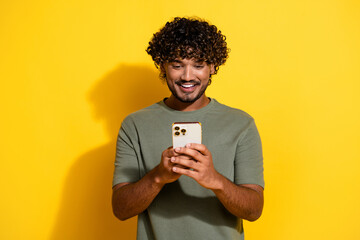 Photo of smart positive guy with wavy hairdo dressed khaki t-shirt look at smartphone typing message isolated on yellow color background