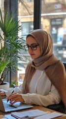 A Muslim woman in a hijab works at her desk with a laptop, writing notes on paper, working online or studying for school