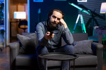 Fototapeta premium Bored person on couch using TV to binge series on subscription based streaming services, browsing using remote. Weary man watching VOD shows on television display, zapping though channels