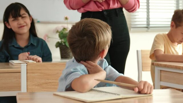Caucasian smart boy raising hand for asking and answering teacher surrounded by diverse children studying at classroom while teacher walking pass at elementary classroom. Education concept. Pedagogy.