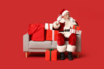 Cool Santa Claus with gift boxes, cookie and glass of milk sitting on sofa against red background