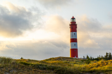 A striking red and white lighthouse stands tall against a colorful sunset sky, surrounded by rolling hills and lush greenery