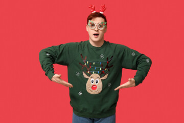 Handsome young man Christmas novelty glasses and reindeer headband showing his warm sweater on red background