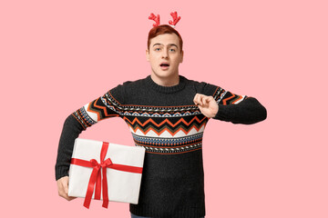 Surprised young man in warm sweater and reindeer headband holding gift box on pink background