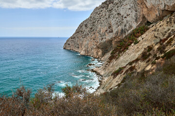 Picturesque view from the top of a cliff with turquoise sea waves meeting the rocky shore, surrounded by lush greenery and a bright sky on a sunny day