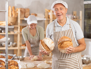 Bakery mature man employee holds ready-made loafs of bread. He stands against background of bakerys...