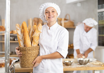 Smiling young female baker holding basket of golden crispy baguettes, proudly presenting freshly...