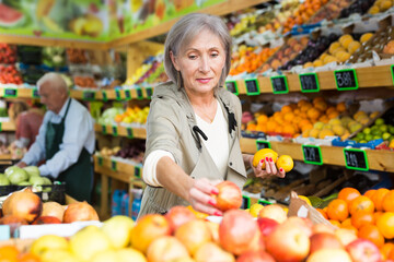 Obraz premium Mature woman standing amongst sheves with fruits and vegetables and selecting fruits. Senior man worker setting out goods in background.