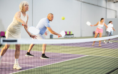Portrait of sporty senior woman playing doubles pickleball with male partner on indoor court, ready...