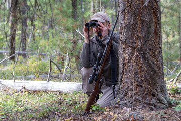 Naklejka premium Hunter hiding behind a tree, watching the surroundings with binoculars. A rifle with the scope is leaning against the tree in front of him.
