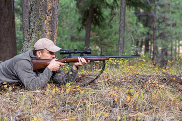 Hunter lying on the ground, aiming his rifle at game while looking through the scope.  Ready to shoot. Side view. Forest on background.