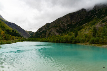 Obraz premium Transparent turquoise river flowing through a lush green valley surrounded by mountains under a cloudy sky