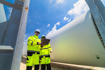 Two engineers in safety gear inspecting a wind turbine blade section on a construction site. They examine the metal framework with precision. Renewable energy project under a clear blue sky.