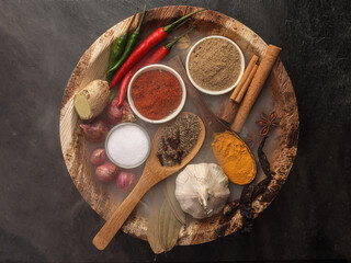 Asian favour spice green red dry chilli powder onion garlic ginger cinnamon cardamon turmeric fennel seeds bay leaf clove star anise wooden spoon blows on palm leaf plate over black slate background