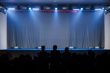 An empty stage illuminated by blue and white spotlights, with a silhouette of the audience seated in the foreground. The atmosphere is set for a performance, show, or event to begin.