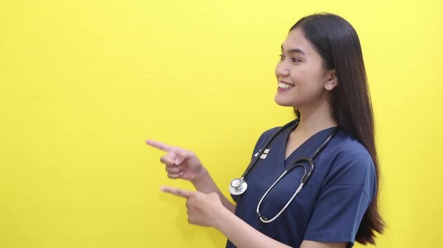 Happy Asian female nurse pointing sideways and showing thumbs up. Isolated on yellow background