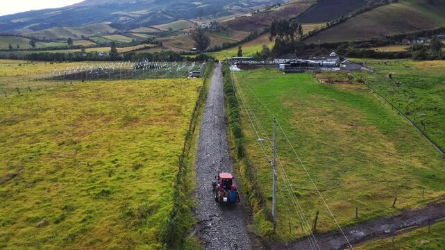 Aerial perspective capturing a tractor traversing a gravel road between vast, lush fields, showcasing the serene and expansive rural landscape with rolling hills in the background under a clear sky