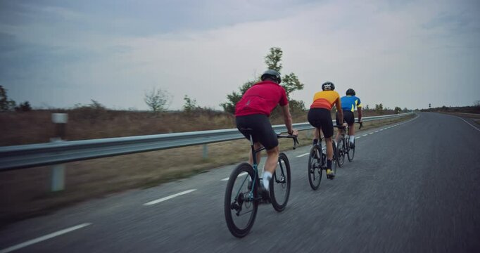 Three Athletes Ride Together Along the Road, Pedaling Bicycles as They Practice on a Country Road. Sportsman in Blue Jersey Leading the Pack and Providing Clean Air Along the Route. Back View