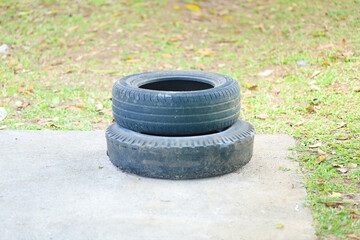 A close-up shot of two old car tires stacked on top of each other, placed on a paved surface outdoors. The background shows a grassy area with scattered dry leaves, indicating an outdoor setting.