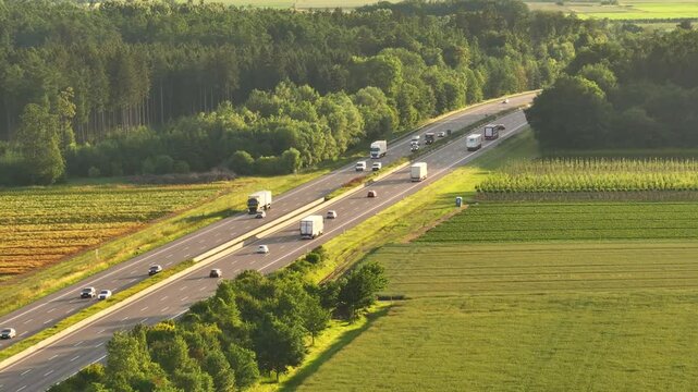 Cars and trucks drive on the A8 highway through lush green fields on a sunny day, aerial view