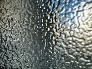 This image shows a close-up of a wavy, textured surface, likely a glass panel with a frosted or patterned design. The light refracts through the glass, creating a distorted, abstract view of the bg