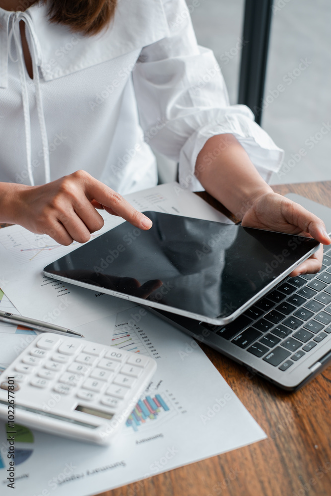 Poster close-up of a business professional using a tablet and laptop for data analysis, surrounded by docum