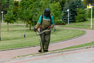 Worker using a grass trimmer to maintain a park, wearing protective gear and cutting grass near a sidewalk