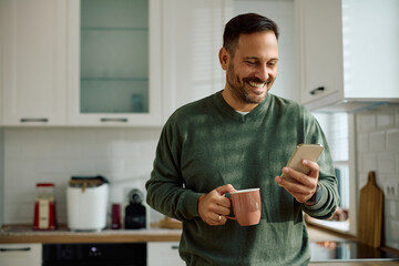 Happy man texting on cell phone while drinking coffee in kitchen.