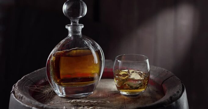 An ice cube slowly falls into a glass, standing on a vintage barrel of whiskey, whiskey from the bottle is immediately poured into the glass. A decanter with a drink stands nearby a dark background