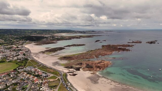 Guernsey Cobo Bay high drone flight showing Cobo village and out to sea over golden beach with rock formations and crystal clear turquoise sea under interesting cloudy sky