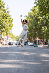 Young woman joyfully jumps in a bustling city street, radiating happiness on a sunny day. Her...