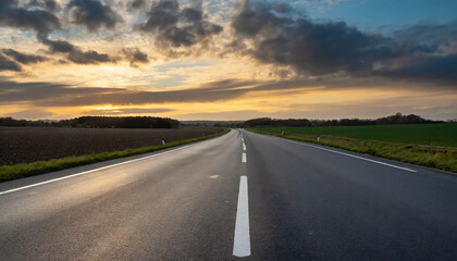 Naklejka premium A peaceful rural road stretches into the horizon beneath a colorful sunset with scattered clouds and lush fields on both sides