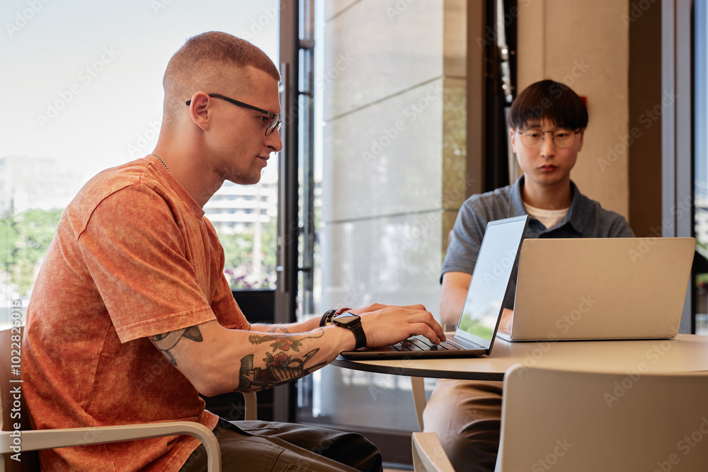 Poster side view of concentrated male it programmer with tattoo on arm wearing glasses to write code on lap