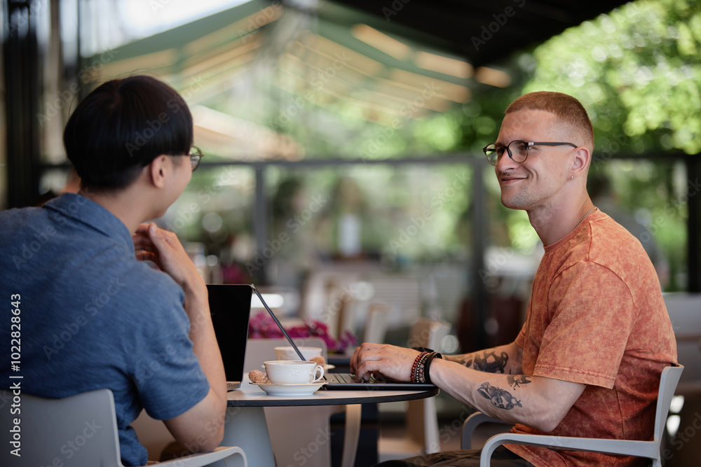 Poster side view of smiling businessman using laptop sitting at cafe table while enjoying talk and collabor