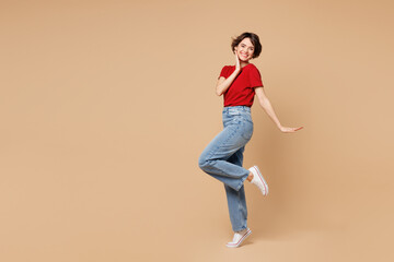 Full body young smiling happy woman she wears red t-shirt casual clothes raise up leg put hand on...