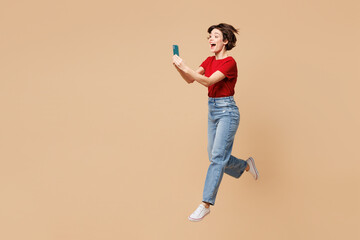 Full body young surprised happy woman wear red t-shirt casual clothes jump high hold in hand use...