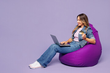 Full body young woman wear denim jacket top casual clothes sit in bag chair use laptop pc computer hold credit bank card shopping online order delivery book tour isolated on plain purple background.