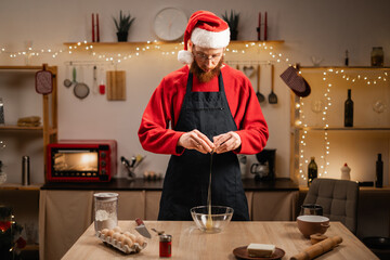 Young man in Santa hat and apron making dough for christmas gingerbread cookies and xmas sweets, bearded guy cooking during winter holidays