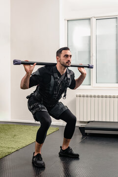 Strong healthy man in special black EMS suit doing squats with bar in sport club, Muscle recovery: Electro stimulator demonstrated by man in club, suitable for fitness recovery visuals