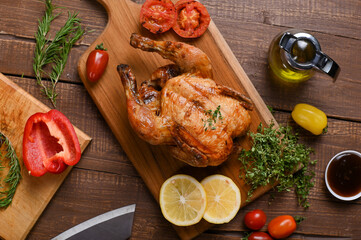 A perfectly roasted whole chicken garnished with herbs on a wooden board, accompanied by sliced lemons, red and yellow bell peppers, grilled tomatoes, fresh rosemary, and olive oil.