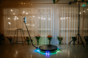 A 360-degree photo booth setup with LED lights and a rotating platform, surrounded by floral...
