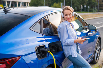 A young woman enjoys music on headphones, leaning against her electric car while it charges.