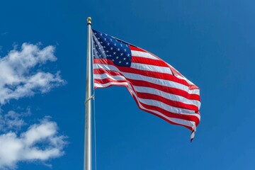 Obraz premium The American flag flutters gracefully atop a tall flagpole, vibrant red and white stripes contrasting with a bright blue sky punctuated by fluffy white clouds.