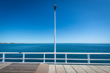 A symmetrical and geometric composition of a pier with a white lamp post and railing against blue sky and water, horizontal
