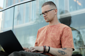 Medium shot of serious male software engineer with septum piercing and glasses typing on laptop while sitting outside leaning against glass construction in city street, copy space